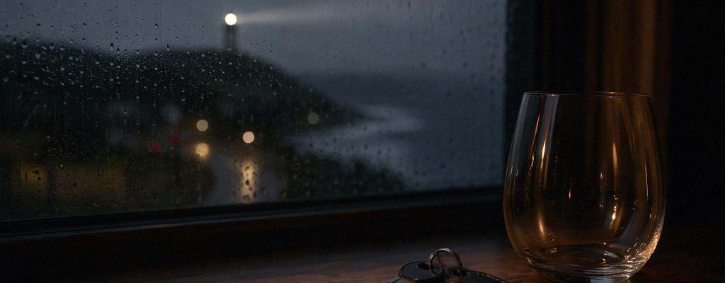 Llaves de coche y vaso de vino vacío sobre mesa oscura junto a ventana con costa atlántica al fondo, evocando un delito de alcoholemia.